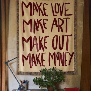 May include: A rectangular woven wall tapestry featuring a beige background with dark red text that reads: MAKE LOVE, MAKE ART, MAKE OUT, MAKE MONEY. The tapestry has a fringed edge and is displayed vertically, showcasing a textured fabric construction.
