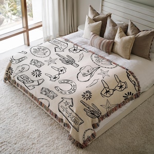 Könnte beinhalten: Eine cremefarbene, gewebte Wohndecke mit einem sich wiederholenden schwarzen Linienmuster aus Western-Motiven, darunter Cowboystiefel, Hüte, Hufeisen, Kakteen, Sterne, Blumen und Wegweiser mit der Aufschrift T-X-T auf einem Bett.