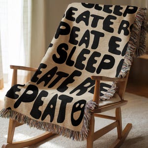 May include: A woven throw blanket with a beige background and black text that reads "SLEEP EAT REPEAT" in a repeating pattern. The blanket features frayed, multi-colored edges and is draped over a wooden rocking chair with a curved backrest.