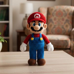 May include: A handmade crochet doll of the video game character Mario. The doll wears a red hat with a white 'M', a red shirt, blue overalls with yellow buttons, and brown shoes. The doll has a tan face, black mustache, and white gloves.