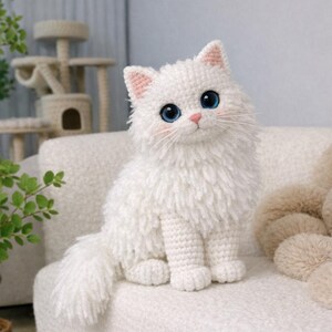 Può includere: Un gatto bianco fatto a mano all'uncinetto, con pelliccia soffice, seduto su un divano bianco. Il gatto ha grandi occhi blu, accenti rosa sulle orecchie e sul naso e lunghi baffi bianchi. Un gatto decorativo carino.