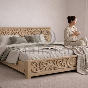 Op de afbeelding: Een licht houten bed met ingewikkelde bloemsnijwerk op het hoofdbord en voetbord. Het bed is opgemaakt met neutraal gekleurd beddengoed en een decoratief kleed. Een persoon zit op het bed.