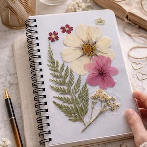 Puede incluir: Un cuaderno con espiral y cubierta azul claro, adornado con flores prensadas en blanco, rosa y rojo. Un bolígrafo dorado descansa junto al cuaderno. El cuaderno está sobre un libro beige. También hay una vela encendida y bolígrafos dorados.