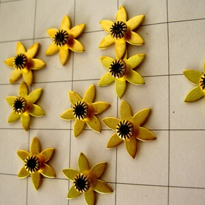May include: Ten gold metal flower-shaped embellishments with black centres. The flowers have five petals and are arranged in a scattered pattern on a white grid background.