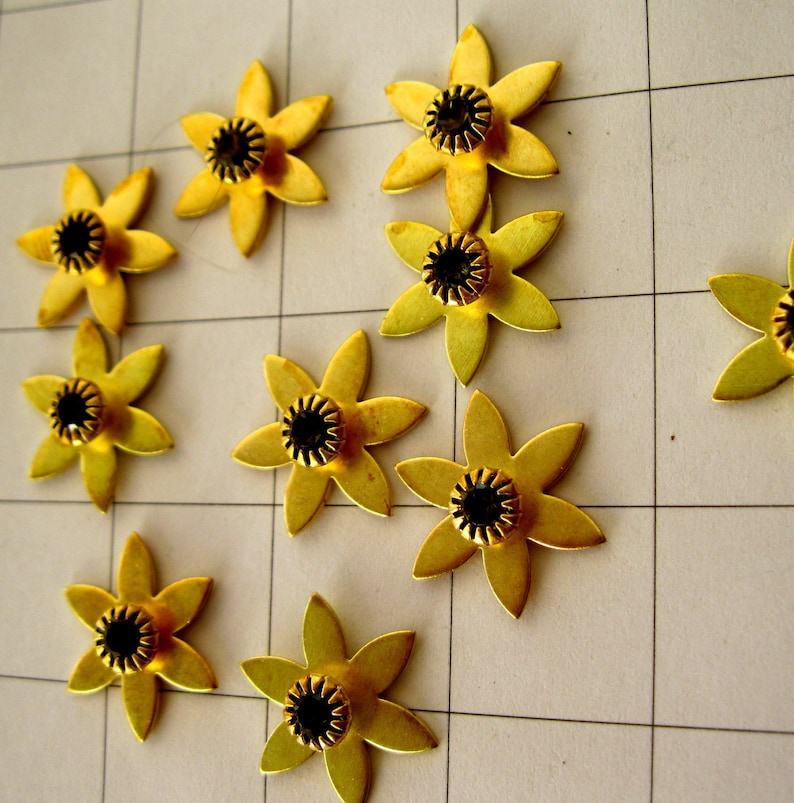 May include: Ten gold metal flower-shaped embellishments with black centres. The flowers have five petals and are arranged in a scattered pattern on a white grid background.