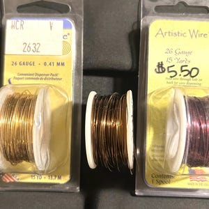 May include: Three spools of Artistic Wire in clear plastic packaging. One spool is gold, one is dark brown, and one is a reddish-purple color. The packaging includes text indicating the gauge, length, and that it is a convenient dispenser pack.