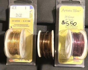 May include: Three spools of Artistic Wire in clear plastic packaging. One spool is gold, one is dark brown, and one is a reddish-purple color. The packaging includes text indicating the gauge, length, and that it is a convenient dispenser pack.