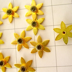 May include: Ten gold metal flower-shaped embellishments with black rhinestones in the centre. The embellishments are arranged on a white grid background.