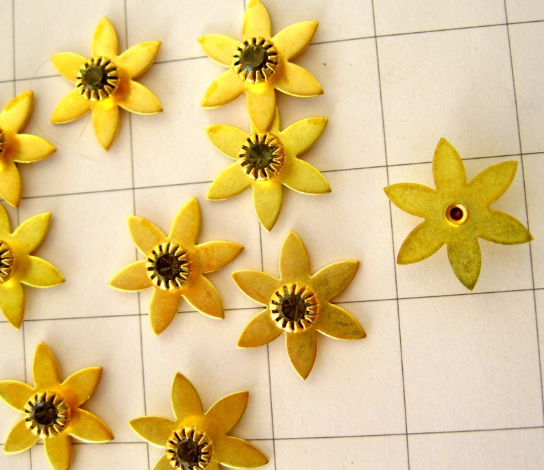 May include: Ten gold metal flower-shaped embellishments with black rhinestones in the centre. The embellishments are arranged on a white grid background.