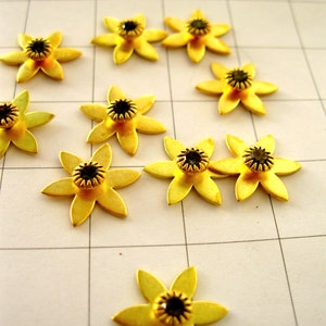 May include: A collection of ten gold-coloured metal flower-shaped embellishments with black rhinestone centres. The flowers are arranged on a white gridded surface.