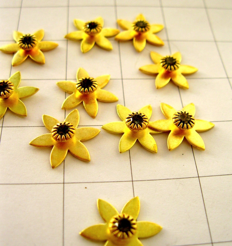 May include: A collection of ten gold-coloured metal flower-shaped embellishments with black rhinestone centres. The flowers are arranged on a white gridded surface.