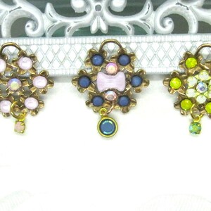 May include: Three gold-tone charms with a flower design. Each charm has a different color scheme: pink, blue, and green. Each charm has a small dangling crystal.