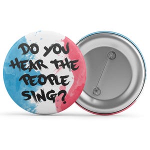 Pode incluir: Um botão redondo com o texto "DO YOU HEAR THE PEOPLE SING?" em preto, sobre um fundo branco com salpicos de aquarela azuis e rosa. A parte de trás do botão é prateada com um alfinete.