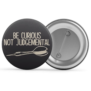 May include: A black button with the text "BE CURIOUS NOT JUDGEMENTAL" in a light beige color, along with a dart illustration. The back of the button is silver with a pin.
