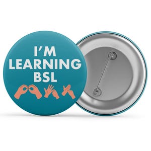 Op de afbeelding: Een turquoise button met de witte tekst "I'M LEARNING BSL". Daaronder illustraties van handen in gebarentaal. De achterkant van de button is zilverkleurig met een veiligheidsspeld.