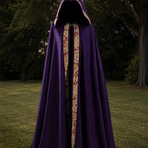 May include: A floor-length deep purple cloak featuring a hood and a front opening trimmed with a wide band of intricate gold and red patterned fabric. The garment is secured at the neck with a decorative metal clasp, creating a draped silhouette.