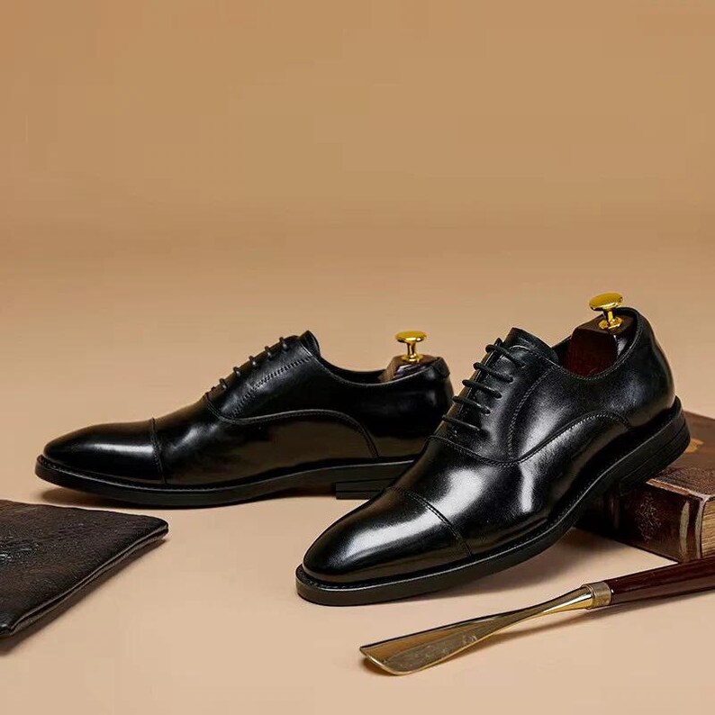 Puede incluir: Un par de zapatos de vestir de cuero negro con dise&ntilde;o de puntera. Los zapatos est&aacute;n atados con cordones y tienen un acabado pulido. Se insertan hormas en los zapatos. Tambi&eacute;n se ven un calzador y una cartera de cuero.