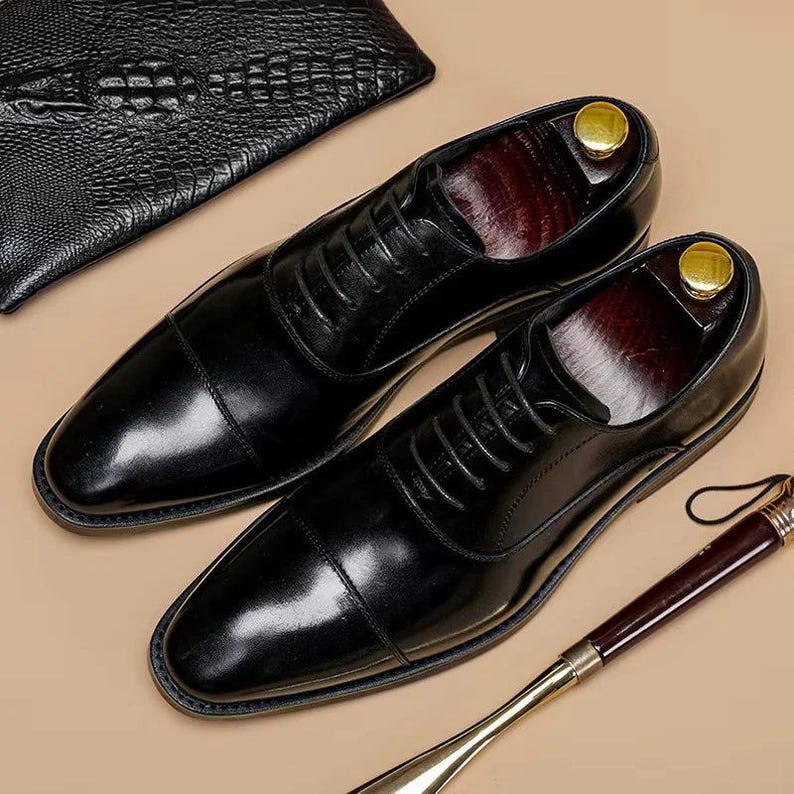 Puede incluir: Un par de zapatos de vestir negros de cuero brillante con un dise&ntilde;o cl&aacute;sico de puntera. Los zapatos tienen cordones y se exhiben con hormas y un calzador. Una cartera de cuero negro est&aacute; en el fondo.