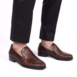 Op de afbeelding: Bruin leren geweven loafers. De schoenen hebben een instapontwerp met een lage hak en een textuur met een mandweefpatroon. De schoenen worden gedragen met een zwarte broek.