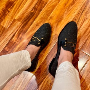 Men’s black glitter loafers with gold horsebit detail styled with cream trousers on wooden floor luxury fashion look.
