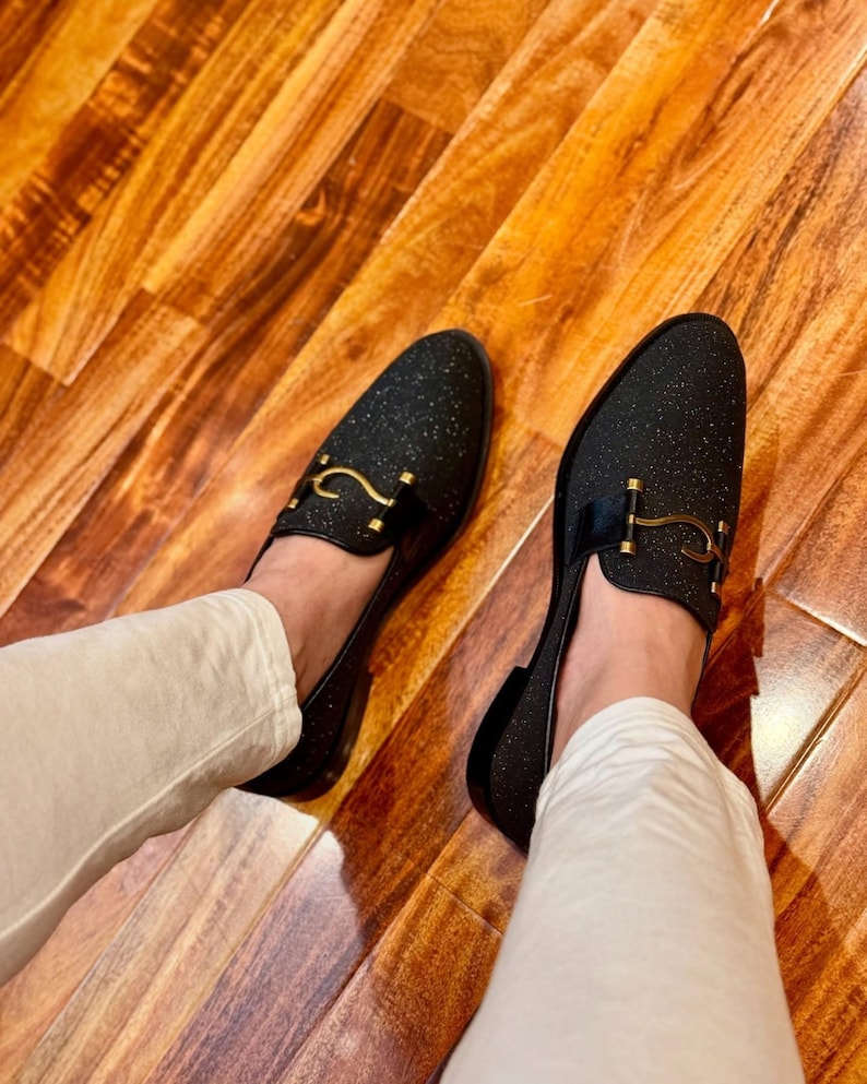 Men’s black glitter loafers with gold horsebit detail styled with cream trousers on wooden floor luxury fashion look.