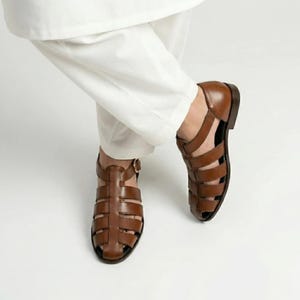 May include: Brown leather sandals with a cage-like design. The sandals have a buckle closure and a low heel. The image shows the sandals being worn with white pants.