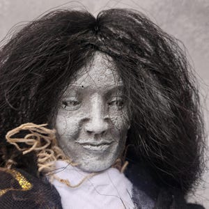 May include: A handmade doll head with a pale, textured face and long, black hair. The doll wears a white shirt and a dark jacket with gold accents. The doll's neck is adorned with twine.
