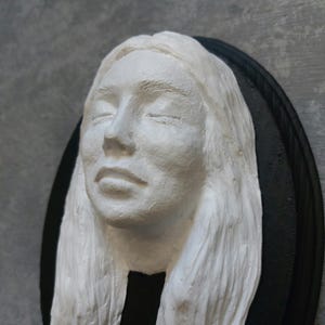 May include: A white plaster sculpture of a woman's face with closed eyes and long hair. The sculpture is mounted on a black circular wooden base.