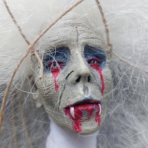 May include: A sculpted doll head with textured gray skin, deep-set eyes with dark blue shading, and red paint dripping from the eyes and mouth. The figure features two prominent white fangs and is surrounded by wispy white hair and thin twine.