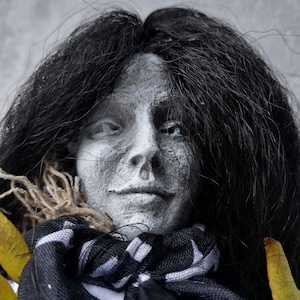 Puede incluir: Primer plano de una cabeza de muñeca con rostro pálido y texturizado y cabello largo y negro. La muñeca lleva una bufanda en blanco y negro y tiene un elemento similar a la paja alrededor del cuello. Tema de Halloween o gótico.