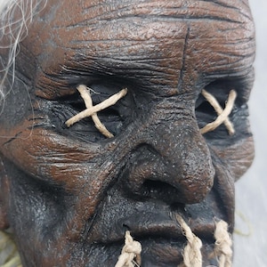 May include: A sculpted brown face with deep wrinkles and a textured surface. The eyes are covered with beige twine stitched in an X pattern. The mouth is sewn shut with three vertical pieces of beige twine. Wispy white hair frames the top of the head.