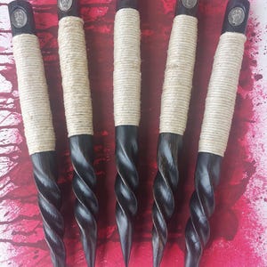 May include: Five dark wooden stakes arranged vertically. Each stake features a spiraled, pointed bottom section and a smooth upper handle wrapped in light beige twine. A small, circular silver-toned metal emblem is embedded at the top of each handle.