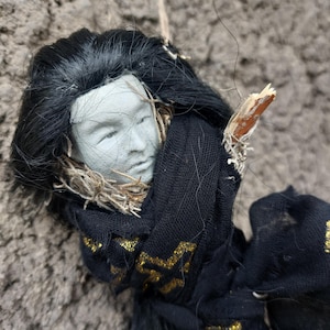 May include: A handmade doll head with black hair and a gray face. The doll is wrapped in black fabric with gold accents. A twig is attached to the doll's head. The doll is hanging against a textured, gray background.