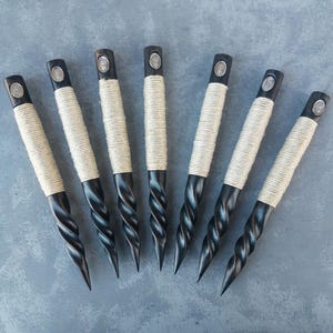 Pode incluir: Oito ferramentas de madeira preta com uma corda branca envolta. As ferramentas têm uma ponta pontiaguda e uma parte superior prateada decorativa.