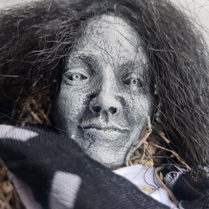 May include: A gray-toned, sculpted face with dark hair, possibly a Halloween decoration. The face has detailed features and is partially obscured by dark hair and a black and white striped fabric.