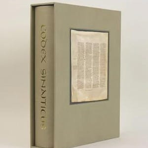 May include: A gray book with the text "CODEX SINAITICUS" in gold lettering on the spine. The book is in a matching gray slipcase. A rectangular window on the front reveals a page of text.