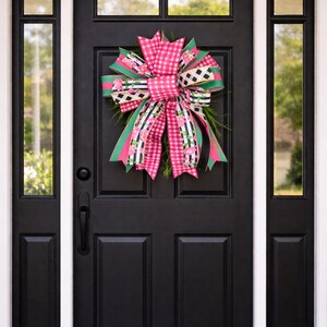 May include: A black front door with a decorative bow. The bow features pink gingham, black and white checkered, and floral patterned ribbons. Greenery accents the bow, creating a welcoming and festive entryway decoration.