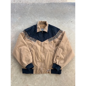 May include: A tan and black jacket with a zip-up front. The jacket has a black yoke and cuffs, with an embroidered geometric pattern across the shoulders. It features a collared neckline and two front pockets. The jacket is made of a durable material.
