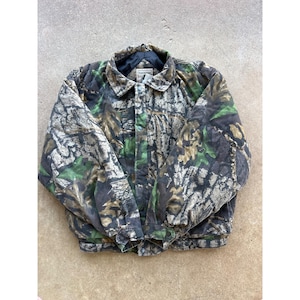 May include: A quilted jacket featuring a forest camouflage pattern in shades of brown, black, and green. The jacket has a fold-down collar, a front snap-button closure, long sleeves with snap-button cuffs, and a black interior lining.