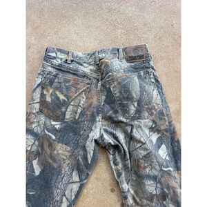 Op de afbeelding: Wrangler jeans met een camouflagepatroon van bruine, grijze en zwarte bosbladeren en boomschors. De broek heeft twee achterzakken, riemlussen en een bruine leren patch op de achterkant van de tailleband met het Wrangler-merklogo.