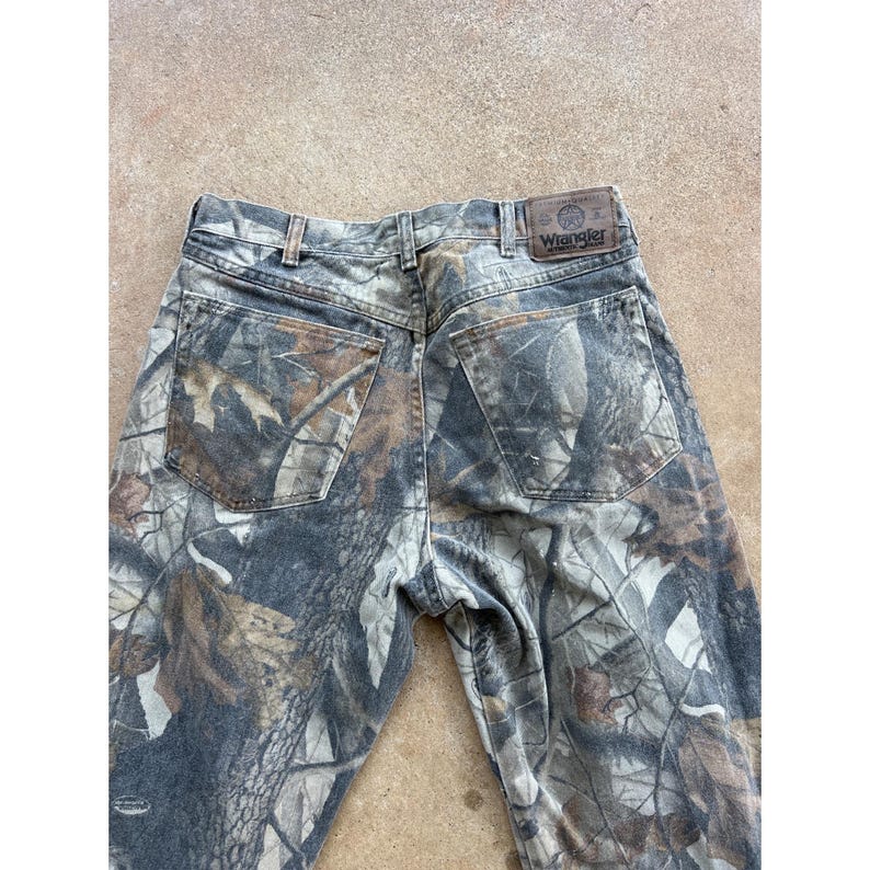Op de afbeelding: Wrangler jeans met een camouflagepatroon van bruine, grijze en zwarte bosbladeren en boomschors. De broek heeft twee achterzakken, riemlussen en een bruine leren patch op de achterkant van de tailleband met het Wrangler-merklogo.