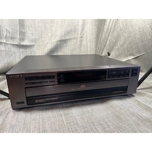 May include: A silver Sony CD player with a black display and control buttons. The player has the Sony logo on the left side and the words "HIGH DENSITY LINEAR CONVERTER" printed on the bottom.