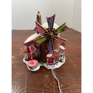 May include: A whimsical Christmas village windmill with a brick tower, colorful blades, and a snow-covered roof. The windmill features a red door, a candy cane, and a sign that says "Candy Cane Lane".