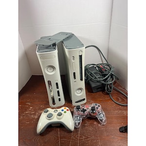 以下が含まれることがあります： 白とグレーのXbox 360コンソール2台、白いコントローラーと透明なコントローラー。コンソールは、電源とケーブルを備えた木製の表面にあります。