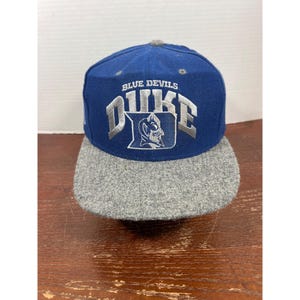 Puede incluir: Una gorra de béisbol azul con visera gris que presenta el texto bordado "BLUE DEVILS" sobre la palabra "DUKE" y un logotipo de una mascota de diablo en hilo blanco y gris en el panel frontal de la corona estructurada.