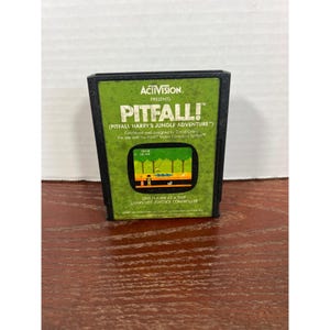 May include: A black Atari 2600 video game cartridge for Pitfall! featuring a green label with the Activision logo, the game title, and a small screenshot of the jungle gameplay. The text includes Pitfall Harry's Jungle Adventure and instructions.