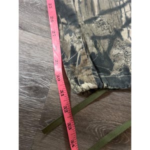 Puede incluir: Pantalones con estampado de camuflaje y cintura con cord&oacute;n. Una cinta m&eacute;trica rosa se coloca junto a los pantalones, mostrando medidas de 63,5 cm a 86,4 cm. Los pantalones presentan colores marr&oacute;n, verde y beige.