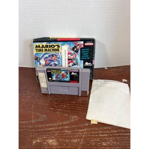 May include: A Super Nintendo Entertainment System video game cartridge for Mario's Time Machine, featuring a gray plastic casing with a colorful label, accompanied by its original cardboard box and a white paper instruction manual.
