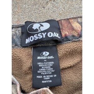 Pode incluir: Close-up de duas etiquetas pretas Mossy Oak. A etiqueta superior tem o logotipo e o texto Mossy Oak. A etiqueta inferior tem o logotipo Mossy Oak, o tamanho XL e detalhes sobre a composi&ccedil;&atilde;o do tecido. O forro &eacute; 100% poli&eacute;ster.