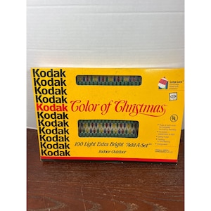 Pode incluir: Uma caixa amarela Kodak de luzes de Natal. A caixa diz "Color of Christmas" e "100 Light Extra Bright 'Add-A-Set' Indoor-Outdoor". A caixa tem um gráfico das luzes e o logotipo da Kodak.